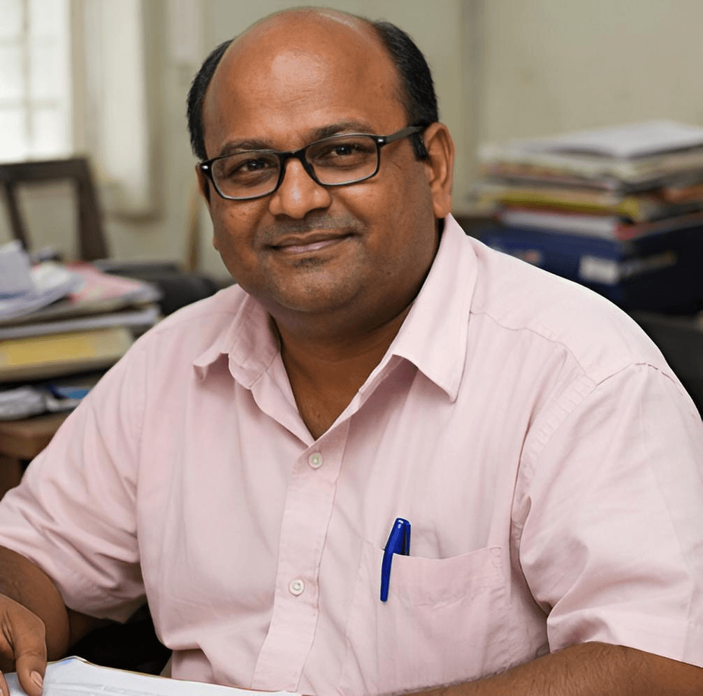 चश्मा और गुलाबी शर्ट पहने एक मुस्कुराता हुआ व्यक्ति अपने ऑफिस डेस्क पर बैठा है।